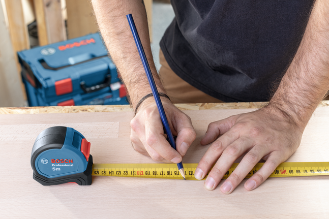 사람이 테이프 측정기와 연필을 이용해 나무에 치수를 표시하고 있습니다.