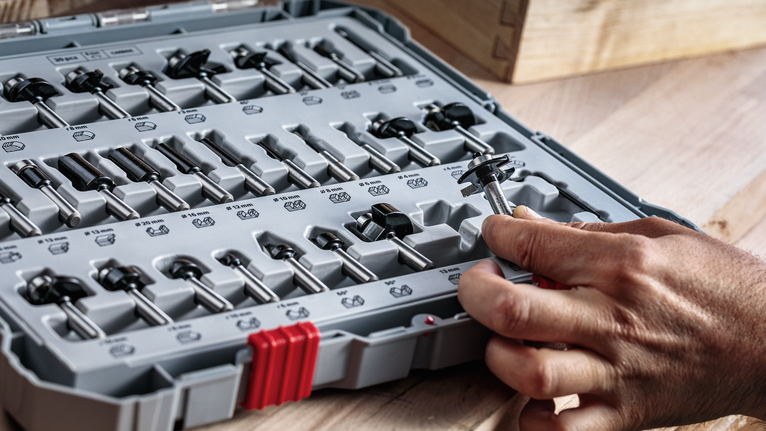 나무 표면 위에 정리된 도구 상자에서 라우터 비트를 선택하는 사람.