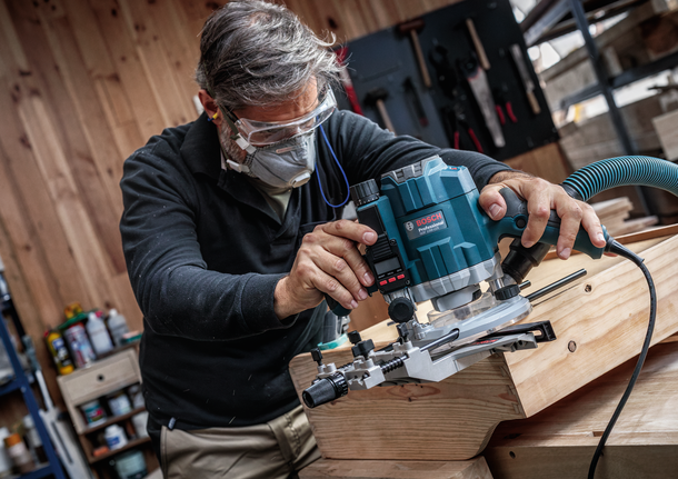 안전장비를 착용한 사람이 라우터를 사용하여 나무판의 가장자리 모양을 만들고 있습니다.