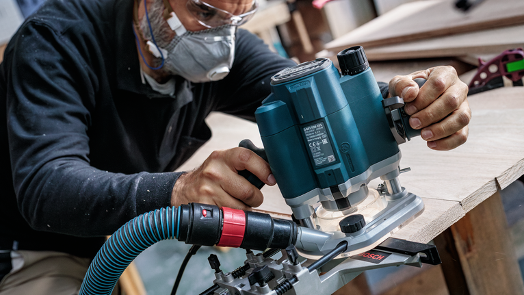 안전 장비를 착용한 사람이 전동 라우터를 사용하여 나무판의 가장자리 모양을 만들고 있습니다.