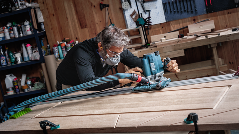 안전 장비를 착용한 사람이 전동 공구를 사용해 나무 문에 홈을 파고 있습니다.