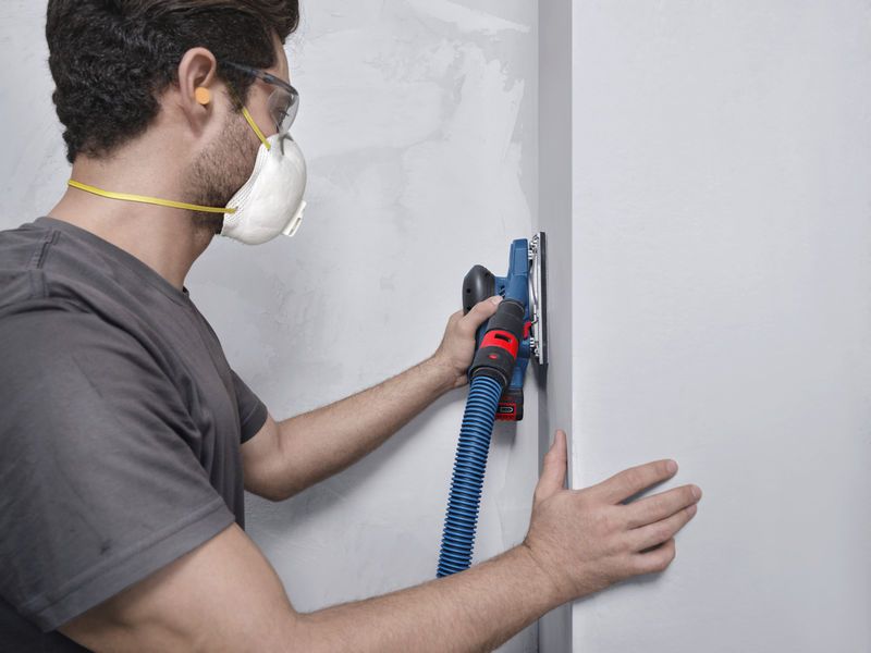 안전 장비를 착용한 사람이 진공 호스에 연결된 궤도 샌더로 벽을 샌딩하고 있습니다.