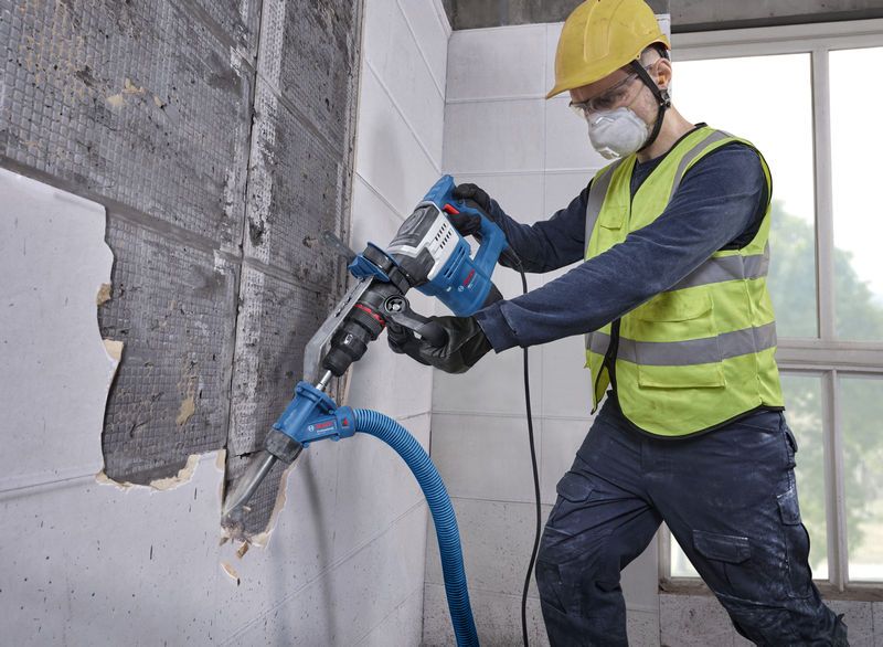 안전 장비를 착용한 사람이 진공 호스가 연결된 전동 공구를 사용하여 벽 타일을 제거하고 있습니다.