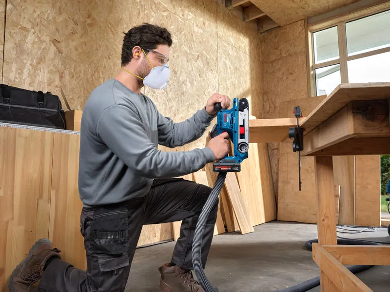 안전 장비를 착용한 사람이 나무판에 벨트 샌더를 사용하고 있습니다.