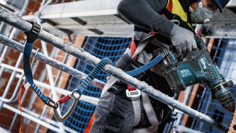 안전장비를 착용한 사람이 비계 위의 콘크리트에 구멍을 뚫고 있습니다.