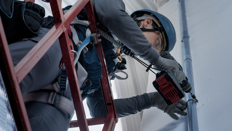 안전장비를 착용한 사람이 전동공구를 사용해 비계에서 벽에 구멍을 뚫고 있습니다.