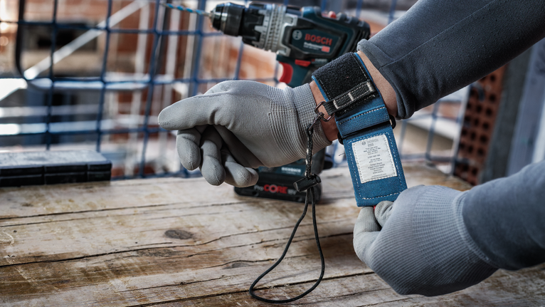 안전 장비를 착용한 사람이 나무 표면 위의 무선 드릴 근처에서 손목 끈을 조정하고 있습니다.