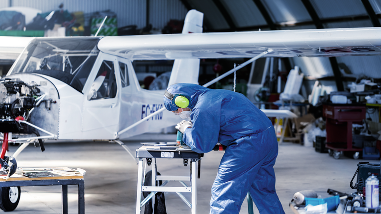 안전 장비를 착용한 사람이 소형 항공기 근처 작업대에서 금속 부품에 구멍을 뚫고 있습니다.