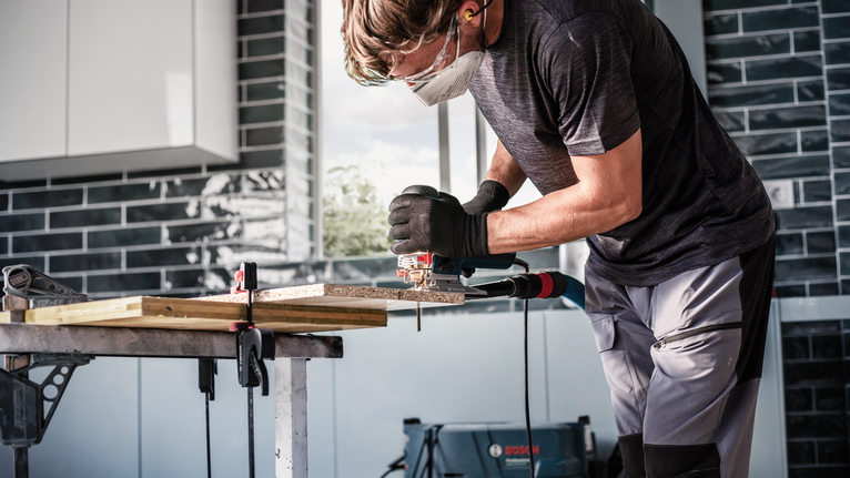 안전 장비를 착용한 사람이 작업대에서 톱을 사용해 나무를 자르고 있습니다.