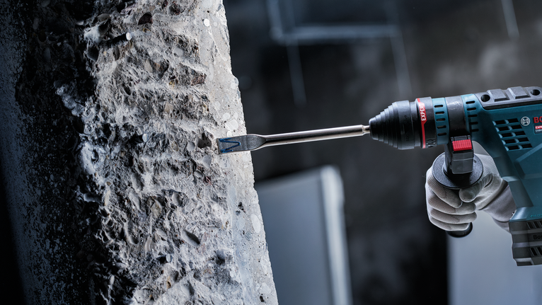 안전 장비를 착용한 사람이 회전 해머 드릴로 콘크리트를 깎고 있습니다.