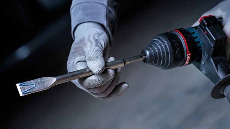 안전 장비를 착용한 사람이 전동 드릴에 끌 비트를 삽입하고 있습니다.