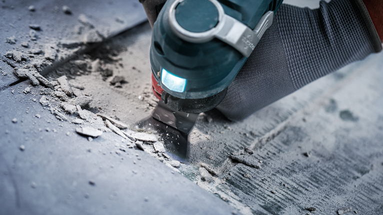 안전 장비를 착용한 사람이 다용도 공구를 사용하여 바닥에서 자재를 제거하고 있습니다.