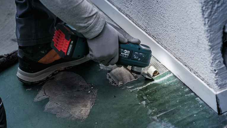 안전 장비를 착용한 사람이 전동 공구를 이용해 벽 근처의 바닥 접착제를 자르고 있습니다.