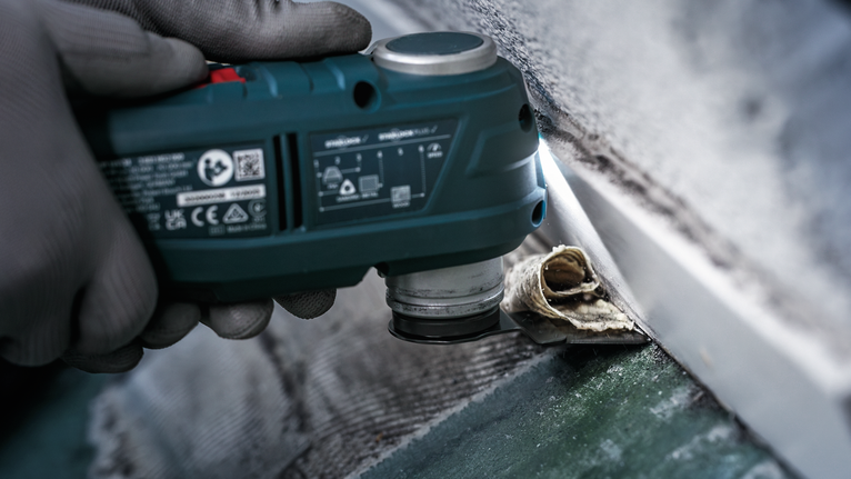 안전 장비를 착용한 사람이 다용도 공구를 사용하여 벽에서 오래된 실리콘 실란트를 제거하고 있습니다.