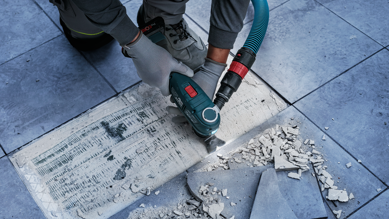 안전 장비를 착용한 사람이 전동 공구를 사용하여 타일 바닥의 타일 접착제를 제거하고 있습니다.