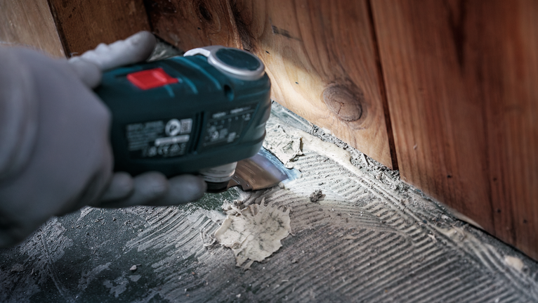 안전 장비를 착용한 사람이 나무 벽 근처 바닥에서 접착제를 긁어내고 있습니다.