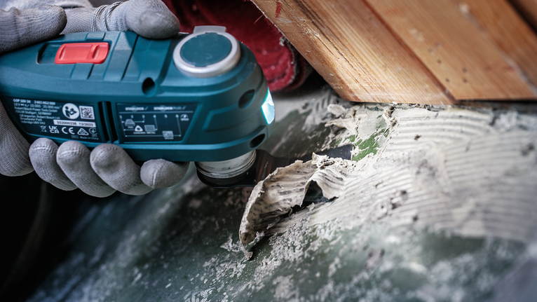 안전 장비를 착용한 사람이 전동 공구를 사용하여 단단한 표면에서 접착제를 제거하고 있습니다.
