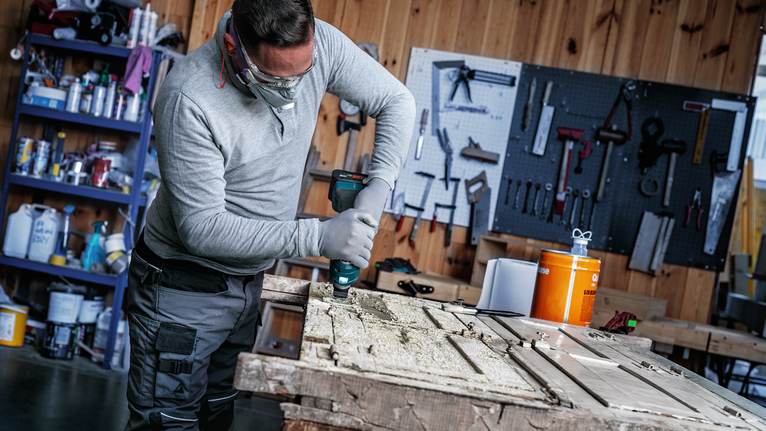 안전 장비를 착용한 사람이 작업대 위의 나무 문에 드릴로 구멍을 뚫고 있다.