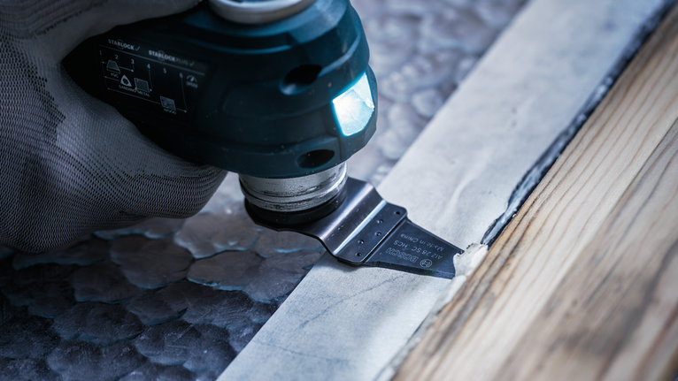 안전 장비를 착용한 사람이 다용도 공구를 사용하여 목재 몰딩 옆의 재료를 자르고 있습니다.