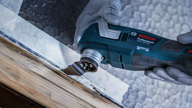 안전 장비를 착용한 사람이 다용도 공구를 사용하여 나무 창틀에서 페인트를 긁어내고 있습니다.