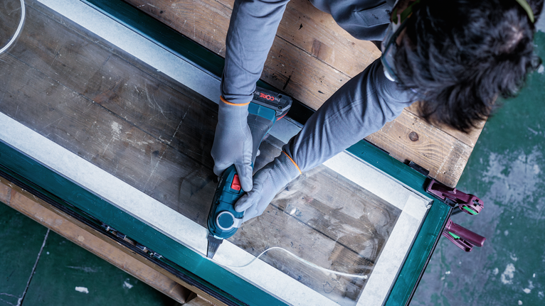 안전 장비를 착용한 사람이 전동 공구를 사용하여 테이프로 표시된 창틀을 따라 절단하고 있습니다.