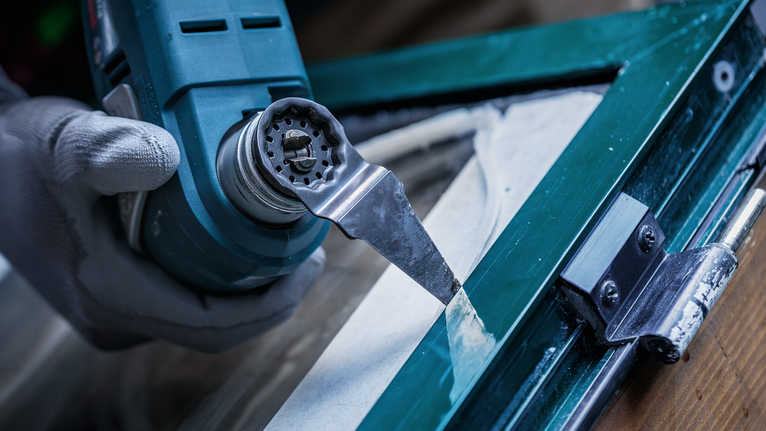 안전 장비를 착용한 사람이 다용도 공구를 사용하여 창문 퍼티를 제거하고 있습니다.