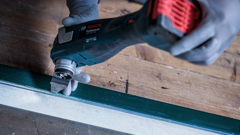 안전 장비를 착용한 사람이 다용도 공구를 사용하여 창틀의 페인트를 사포질하고 있습니다.