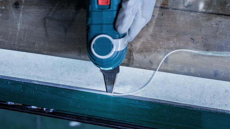 안전 장비를 착용한 사람이 전동 공구를 사용하여 창문 가장자리를 따라 실리콘 실란트를 자르고 있습니다.