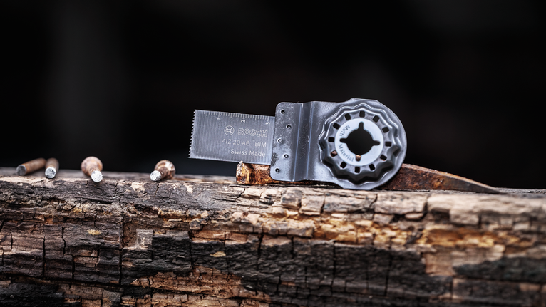 진동식 다용도 공구 날이 낡은 나무 기둥 위에 놓여 있고, 그 옆에는 못 네 개가 박혀 있다.