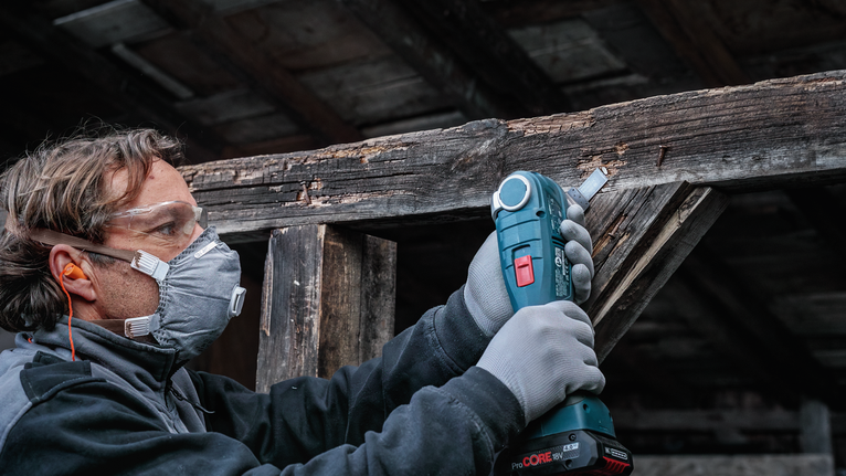 안전 장비를 착용한 사람이 무선 공구를 사용하여 풍화된 나무를 사포질하고 있습니다.