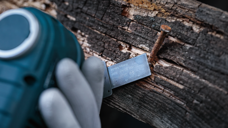 안전 장비를 착용한 사람이 전동 공구를 이용해 나무와 못을 자르고 있습니다.