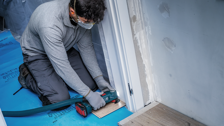 안전 장비를 착용한 사람이 전동 공구를 사용하여 출입구 근처의 바닥재를 자르고 있습니다.