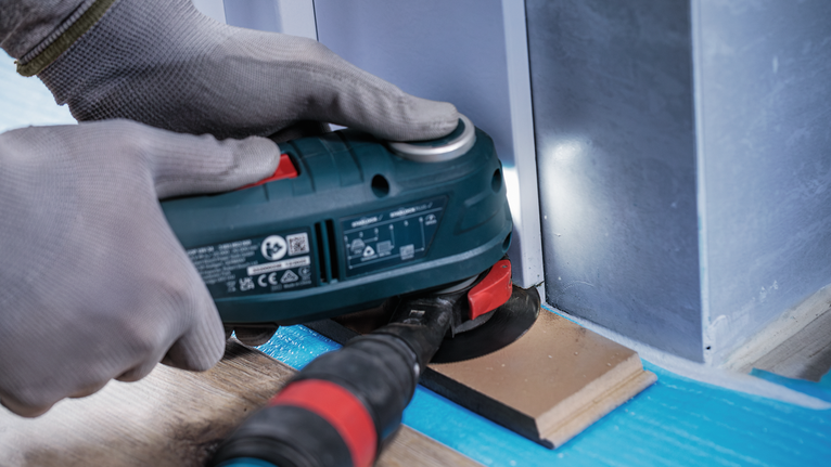 안전 장비를 착용한 사람이 전동 공구를 사용하여 벽 근처의 나무를 사포질하고 있습니다.