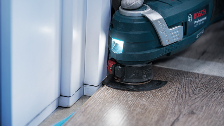 진동식 다용도 공구를 사용하여 문틀 근처의 라미네이트 바닥재를 다듬고 있습니다.