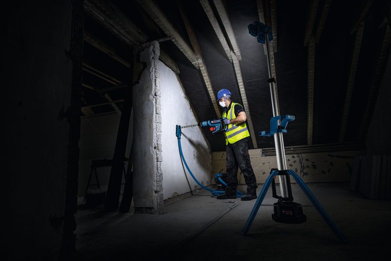 안전 장비를 착용한 사람이 무선 작업 현장 조명으로 밝혀진 벽에 구멍을 뚫고 있습니다.