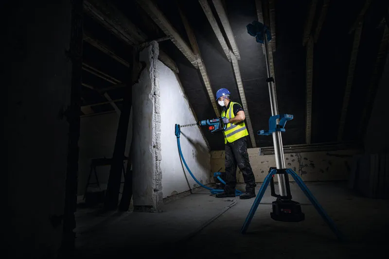 안전 장비를 착용한 사람이 무선 작업 현장 조명으로 밝혀진 벽에 구멍을 뚫고 있습니다.