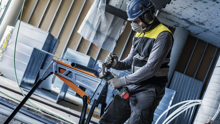 안전 장비를 착용한 사람이 작업대 위의 금속 튜브에 구멍을 뚫고 있습니다.