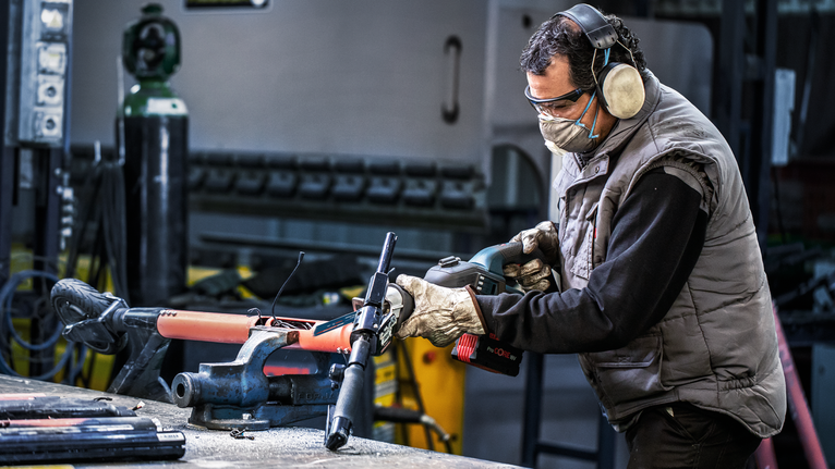 안전 장비를 착용한 사람이 고정된 금속 파이프에 전동 공구를 작동시키고 있습니다.