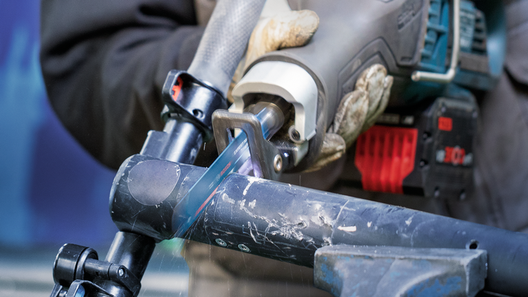 안전 장비를 착용한 사람이 왕복톱을 사용하여 금속 파이프를 절단하고 있습니다.