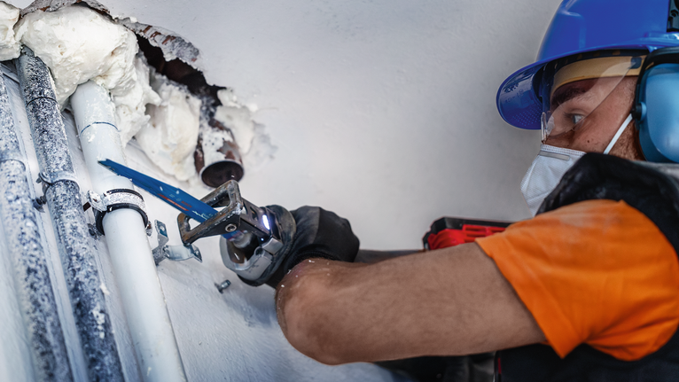 안전 장비를 착용한 사람이 왕복톱을 사용하여 벽 근처의 파이프를 절단하고 있습니다.