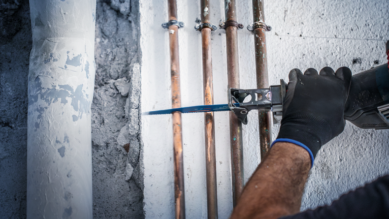 안전 장비를 착용한 사람이 왕복톱으로 배관 근처 벽을 자르고 있습니다.