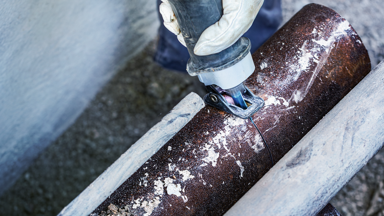 안전 장비를 착용한 사람이 왕복톱을 사용하여 큰 금속 파이프를 자르고 있습니다.