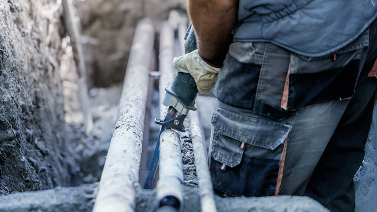 안전 장비를 착용한 사람이 왕복톱으로 도랑 속의 큰 파이프를 자르고 있다.