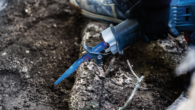 안전 장비를 착용한 사람이 왕복톱으로 두꺼운 나무뿌리를 자르고 있습니다.