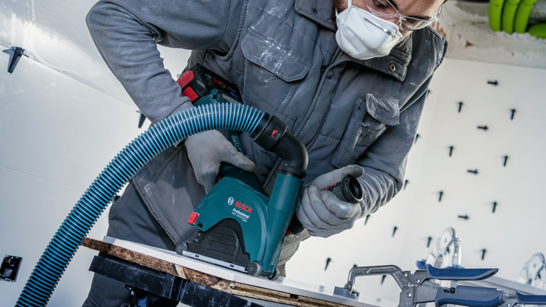 안전 장비를 착용한 사람이 집진 장치가 장착된 전동톱을 사용하여 판자를 자르고 있습니다.