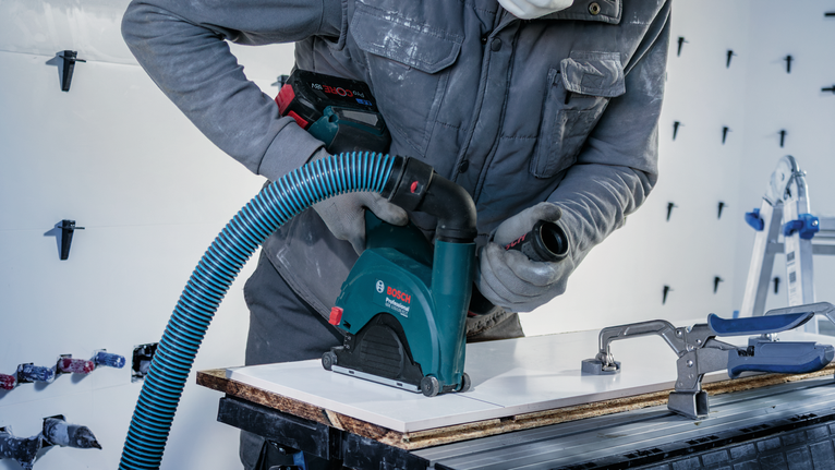 안전 장비를 착용한 사람이 먼지 제거 기능이 있는 전동 공구를 사용하여 타일을 작업하고 있습니다.