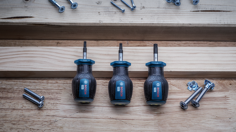 나무판자 위에 드라이버 세 개가 놓여 있고, 그 옆에는 나사와 볼트가 흩어져 있다.