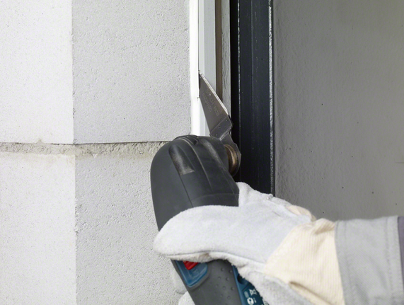 안전 장비를 착용한 작업자가 전동 공구를 이용해 창틀에서 실런트를 잘라냅니다.