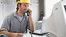 노란 헬멧을 쓴 남성이 컴퓨터 앞에서 전화 통화 중이다.