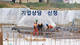 건설 현장에서 작업자들이 기초 공사를 진행하고 있으며, 배경에는 큰 현수막이 걸려 있다.